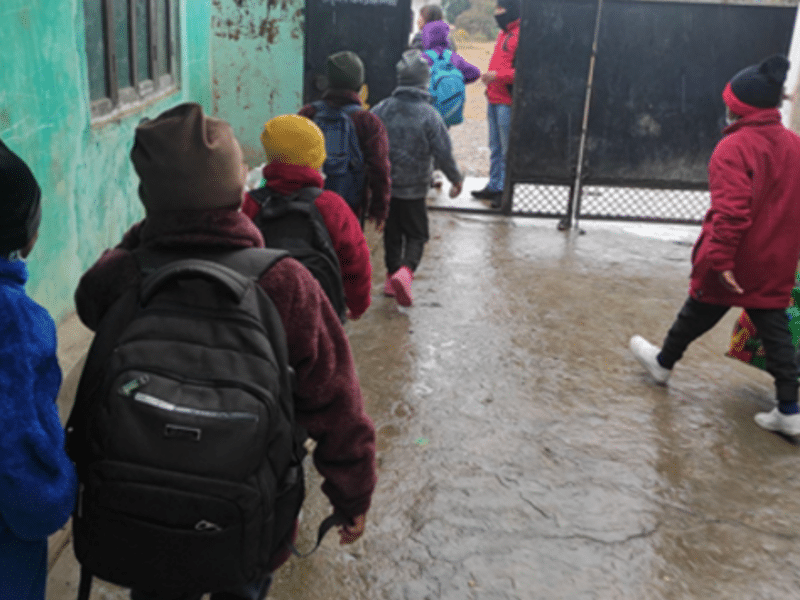 Several warmly dressed Nepali children troop out through an outside door of an orphanage, backs to the camera, wearing rucksacks