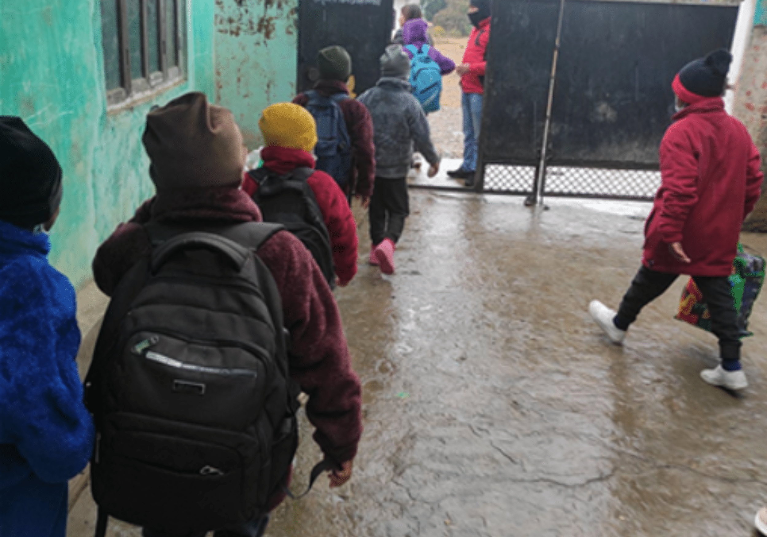 Several warmly dressed Nepali children troop out through an outside door of an orphanage, backs to the camera, wearing rucksacks