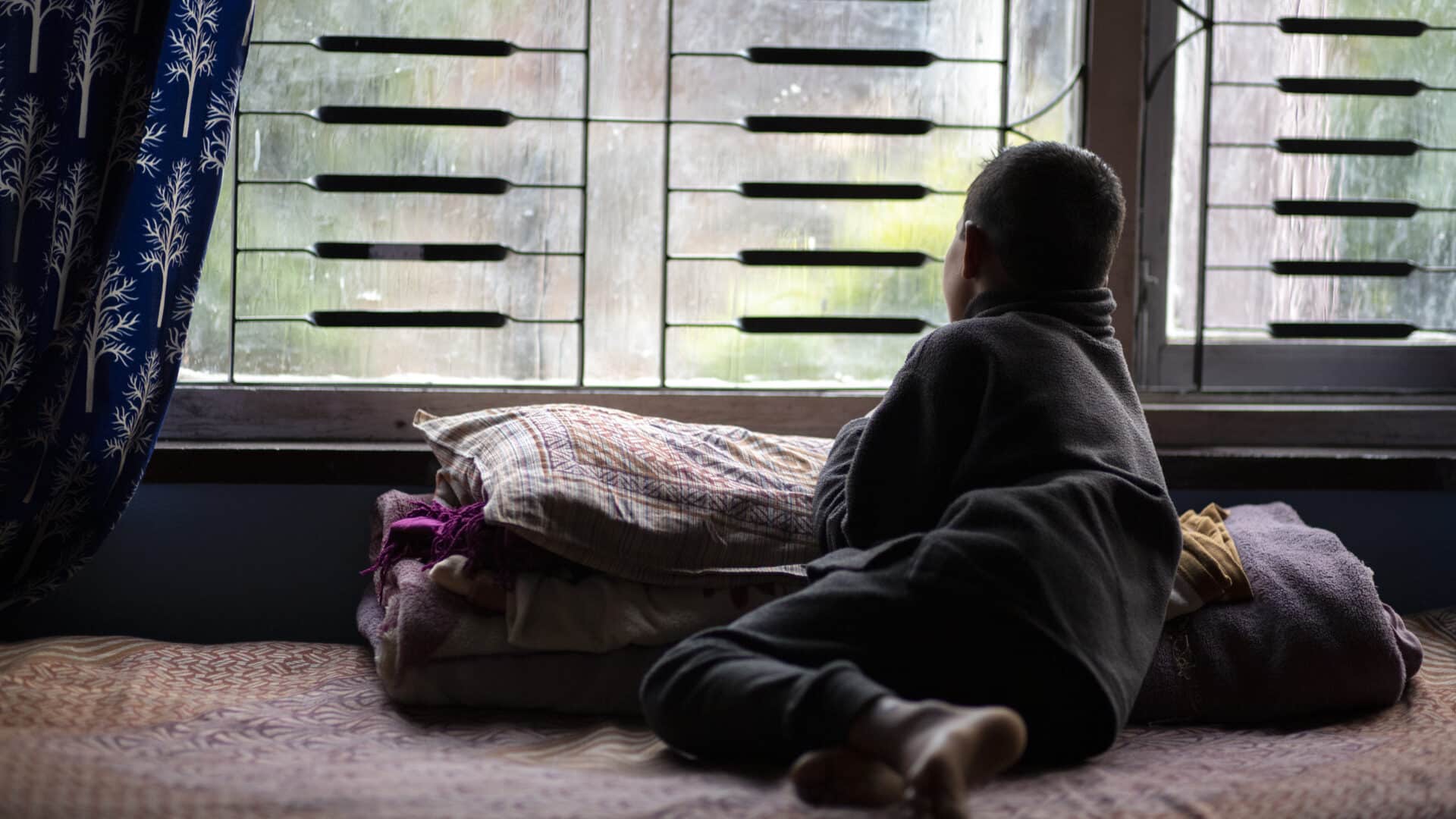 A nepalese child with their back turned gazes sadly out of the barred windows of an orphanage