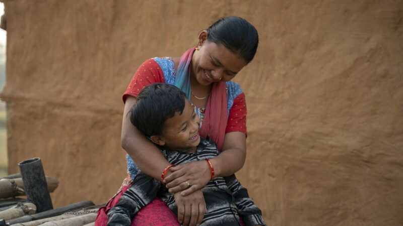 Nepali mother Lata* hugs her 8 year old son Sunil* in front of their red earth wall, after he returns from years in an orphanage