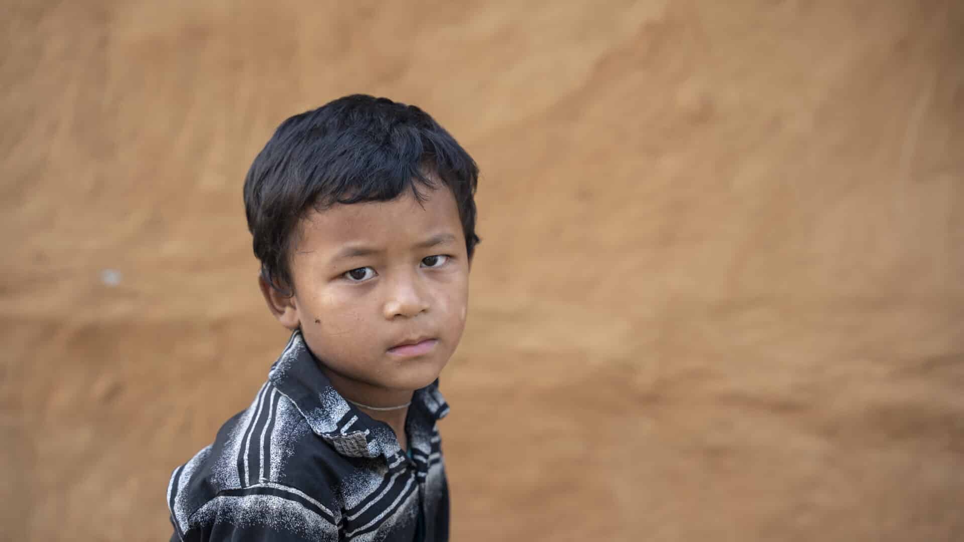 Young Nepali boy with short dark hair, in a striped polo shirt looks sadly sideways towards the camera, in front of brown earthen wall