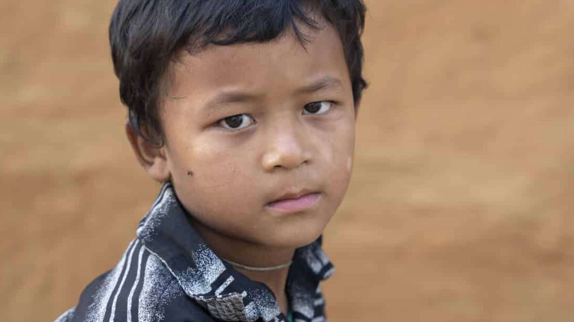 Young Nepali boy with short dark hair, in a striped polo shirt looks sadly sideways towards the camera, in front of brown earthen wall