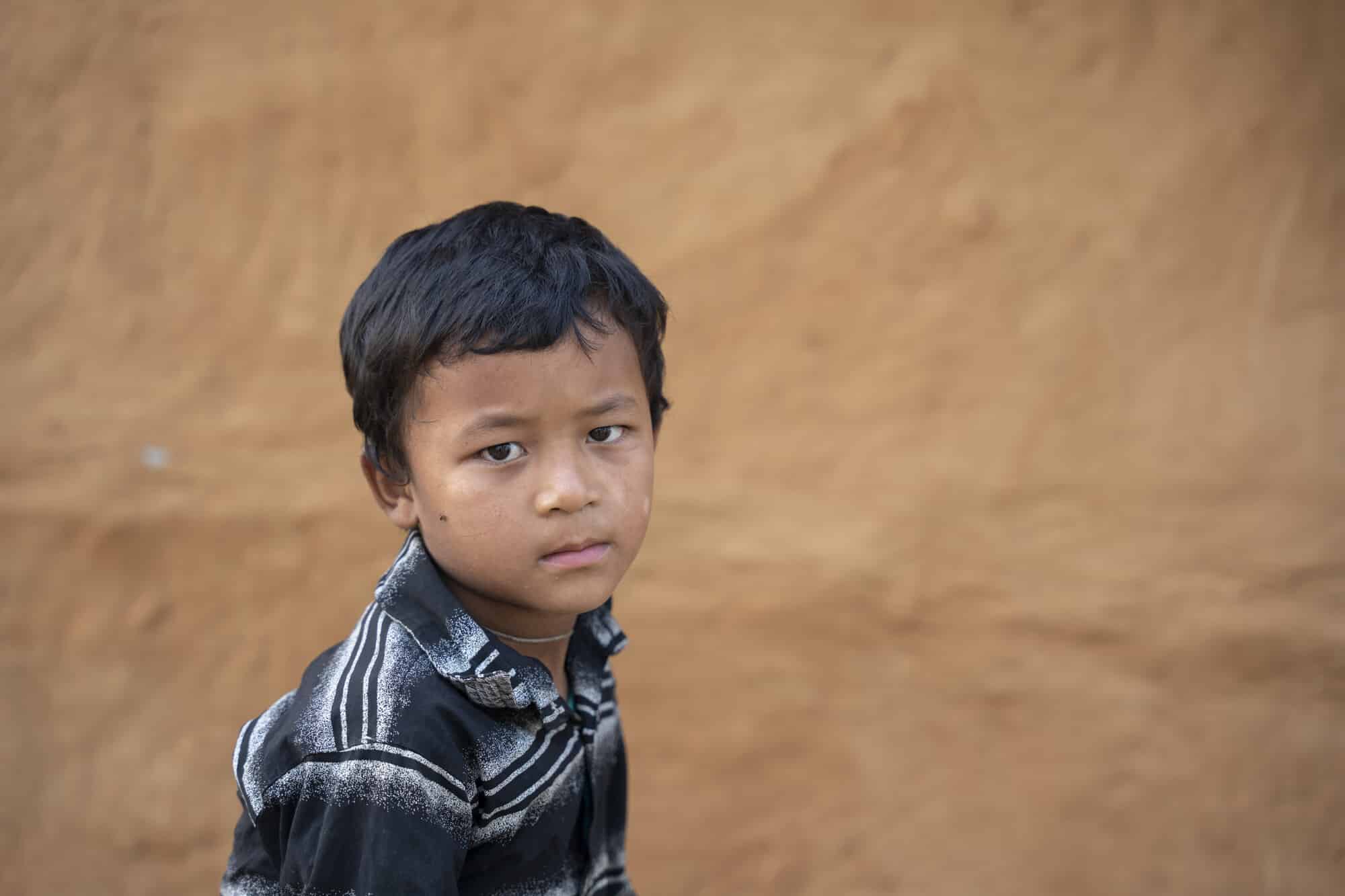 Young Nepali boy with short dark hair, in a striped polo shirt looks sadly sideways towards the camera, in front of brown earthen wall