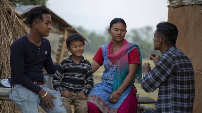 Nepali reintegration officer Rohan talks to a family in a remote district outside their house. The parents look serious while the young son in the middle smiles at Rohan