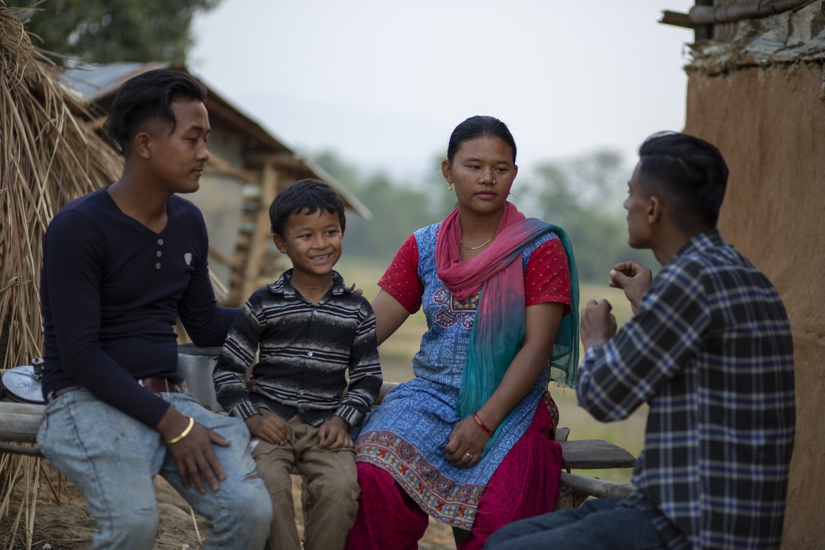 Nepali reintegration officer Rohan talks to a family in a remote district outside their house. The parents look serious while the young son in the middle smiles at Rohan