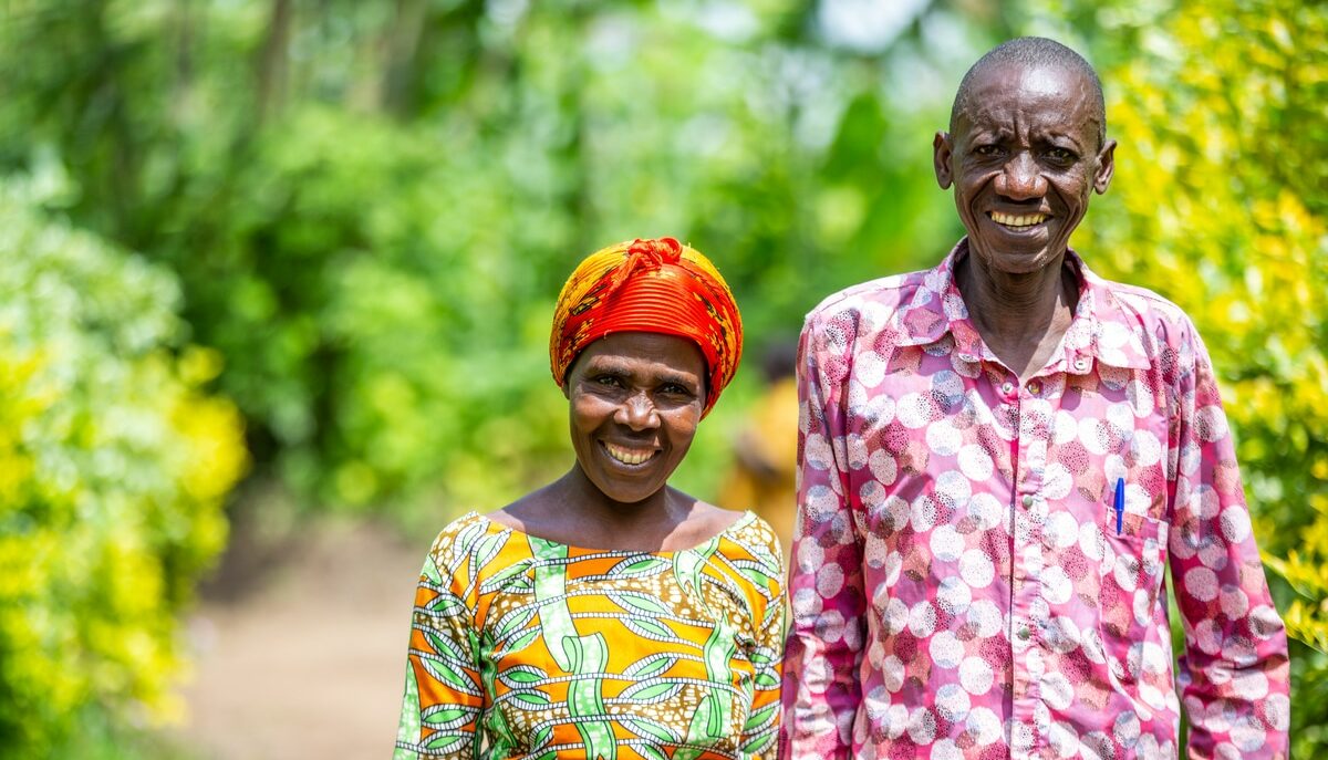 Two older Rwandan foster parents Masaro* and her husband Rugwiro* stand smiling in bright patterned shirts in front of greenery. They look proud and united.