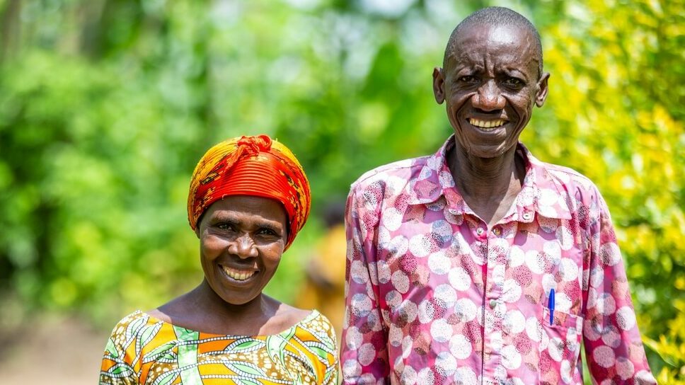 Two older Rwandan foster parents Masaro* and her husband Rugwiro* stand smiling in bright patterned shirts in front of greenery. They look proud and united.