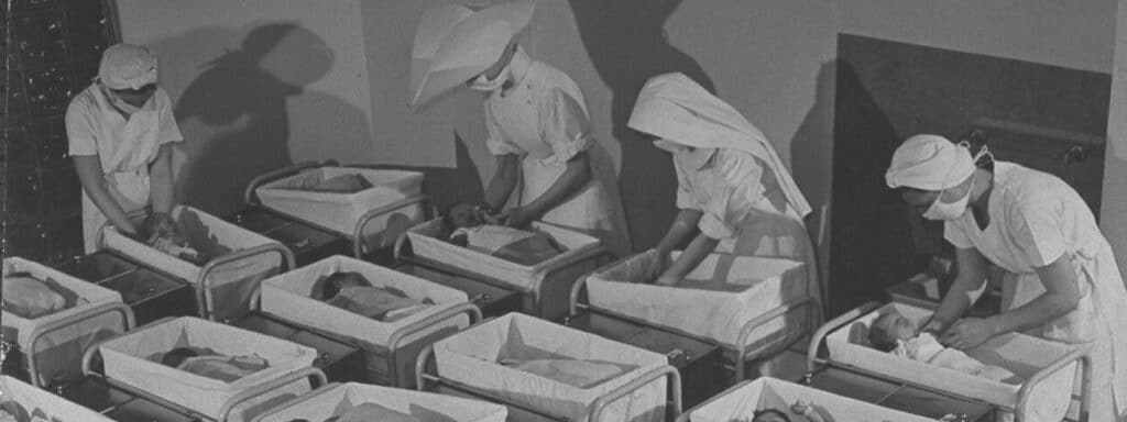 Black and white photo from 1940s or 50s north america of 4 nurses in starched white uniforms and masks tending to babies in several rows of cots in a clinical setting. It feels institutional and cold and there is a lack of parental love or individual care.