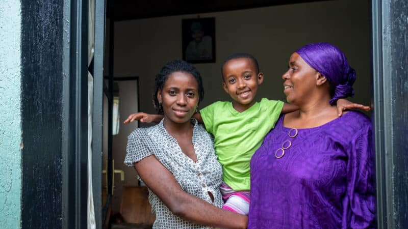A young Rwandan woman with short dark hair and a light blue shirt stands in the doorway holding up her 6 year old daughter, wearing a green t shirt. They're with a larger, older Rwandan lady in a purple top with matching headscarf. They look calm and happy, smiling at the camera and each other.