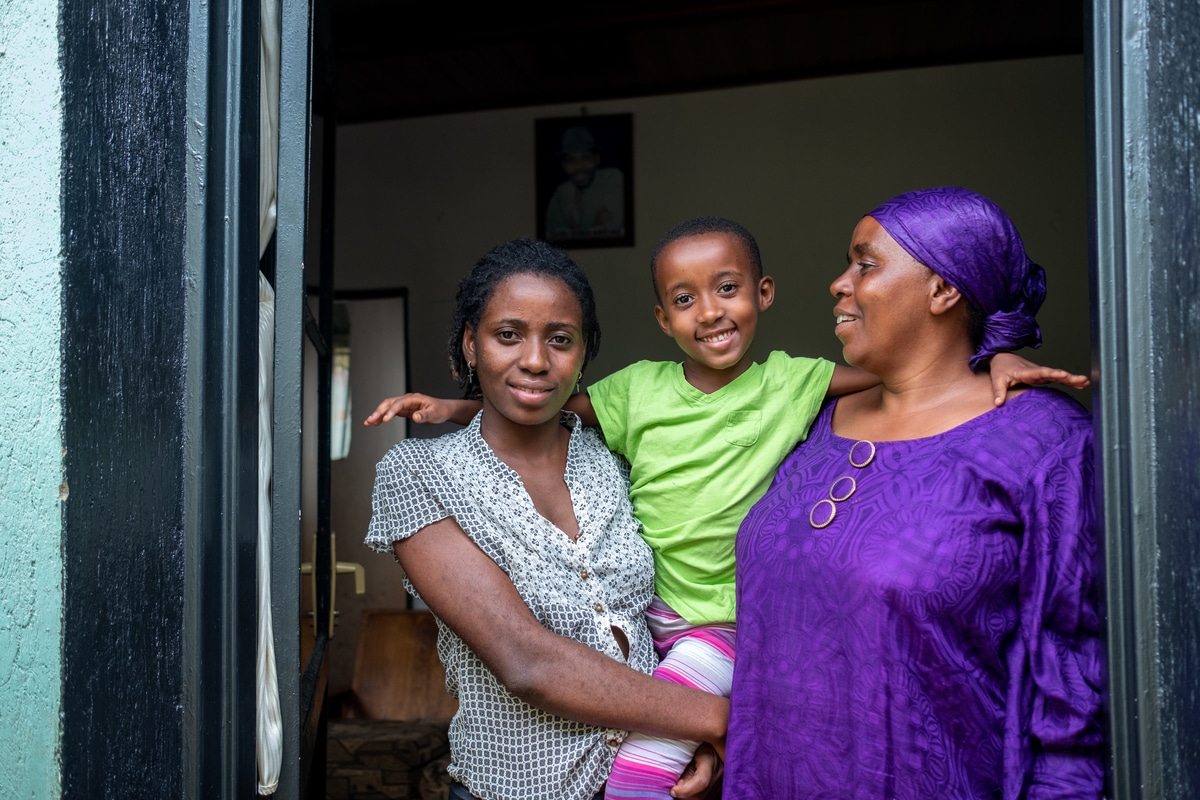A young Rwandan woman with short dark hair and a light blue shirt stands in the doorway holding up her 6 year old daughter, wearing a green t shirt. They're with a larger, older Rwandan lady in a purple top with matching headscarf. They look calm and happy, smiling at the camera and each other.