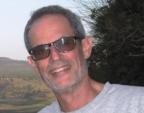 Child development expert Barry Bogin, smiles at the camera, in a close up in front of a green hill and tree. He is an older middle aged white man with short grey hair and stubble, dressed in a light grey t shirt and dark glasses. He looks happy and relaxed.