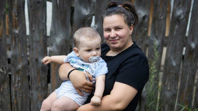 Young brunette mum from Ukraine smiles proudly while holding her baby boy in her arms.