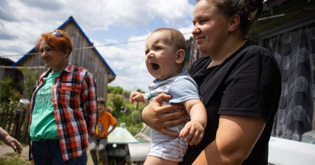 Tina, a young mum, holds her baby boy Serhii. against a blue sky and green meadows in the background.