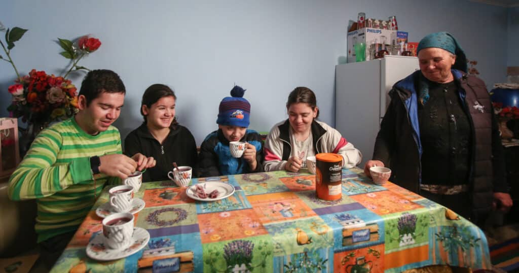 Iulian and Mihaela, home with their siblings, Valeria and Mihai, and their grandma, Gabriela. They sit around a table, sipping coffee and smiling.
