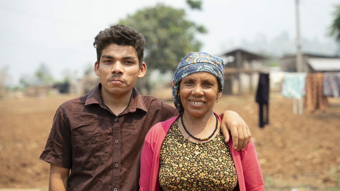Moti* and his mum, Kumari*, from Nepal. They stand outside with a brown field in the background, smiling proudly. Moti has his arm over his mum's shoulders.