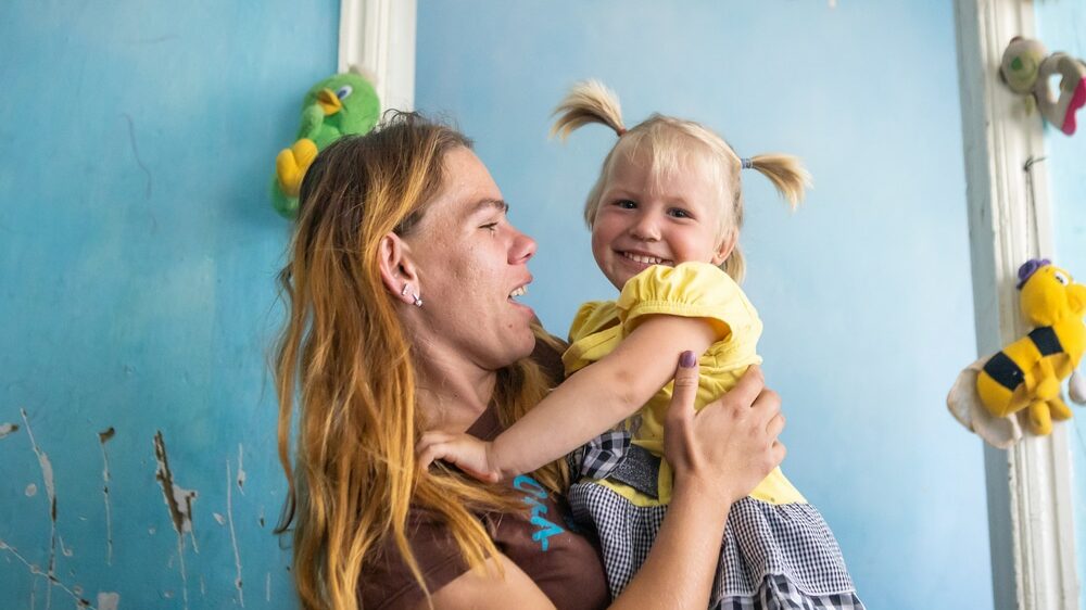 Viktoriia* and her young daughter, in Ukraine. Viktoria is holding her up and smiling at her with a great sense of loving. Her daughter grins at the camera, delighted to be held by her mum.